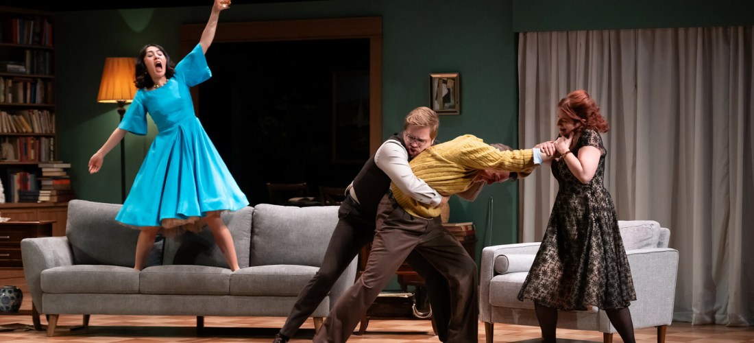 Photo of four actors: three engaged in a struggle, one standing on a couch hoisting a drink and yelling