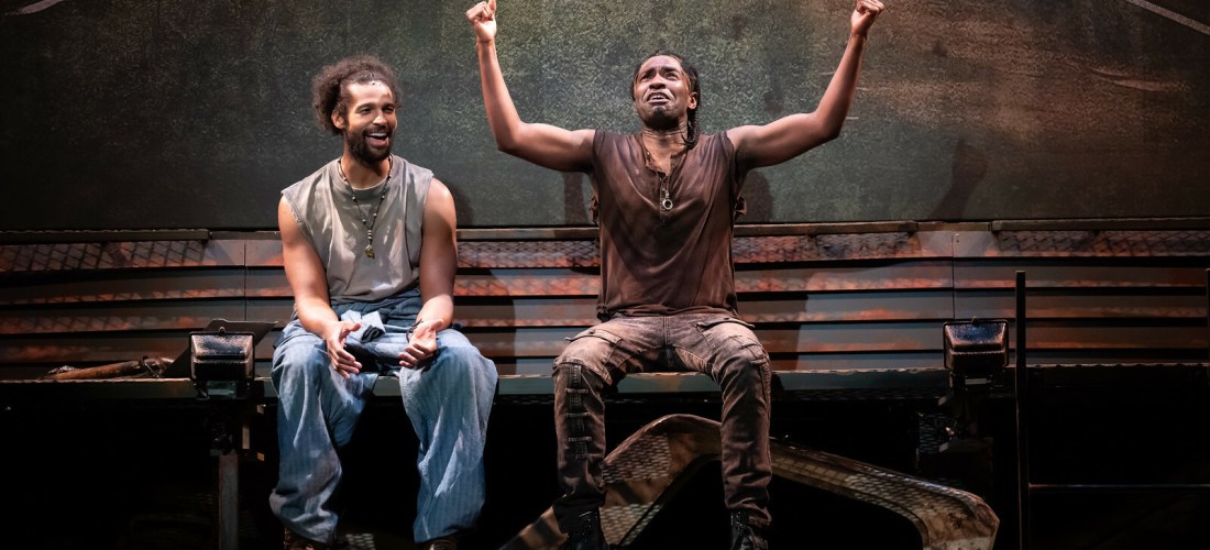 Two actors sit on a platform, one animated with arms in the sky.