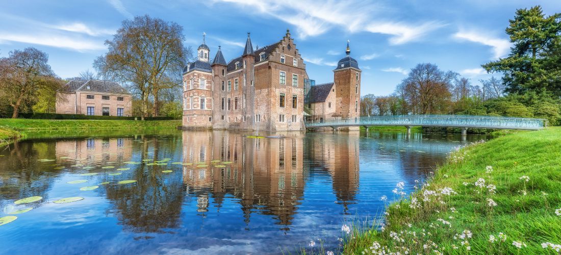 Castle with reflection in a lake surrounded by nature under a bright blue sky.